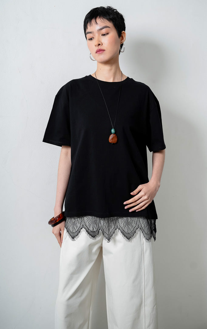 kaos wanita lengan pendek warna hitam (black T-shirt) Lyora Top dengan siluet santai (relaxed silhouette), kerah bulat, dan detail aksen hiasan renda halus di bagian kelim bawah (delicate lace hem detail). 