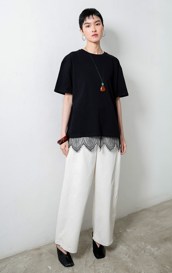 kaos wanita lengan pendek warna hitam (black T-shirt) Lyora Top dengan siluet santai (relaxed silhouette), kerah bulat, dan detail aksen hiasan renda halus di bagian kelim bawah (delicate lace hem detail). 