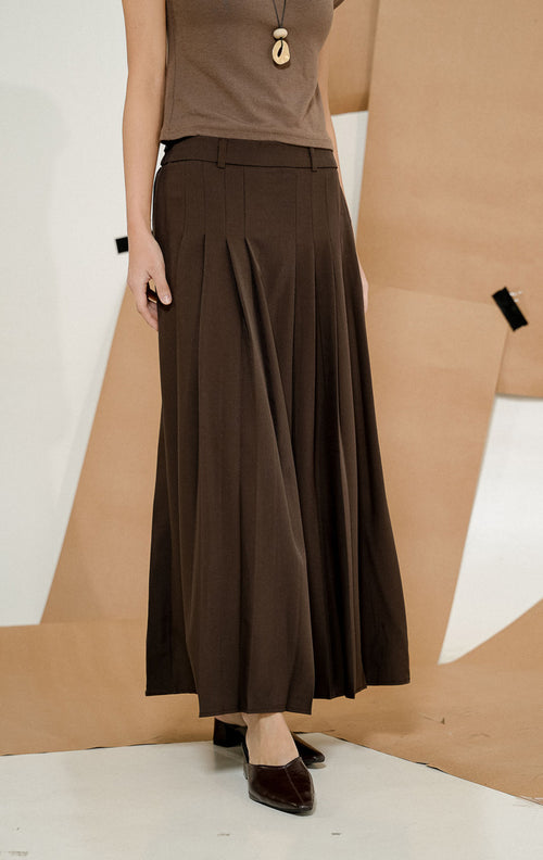 Model berdiri mengenakan Maiden Box Pleats Maxi Skirt Dark Brown, produk rok panjang wanita model maxi warna cokelat tua dengan detail lipit kotak. 