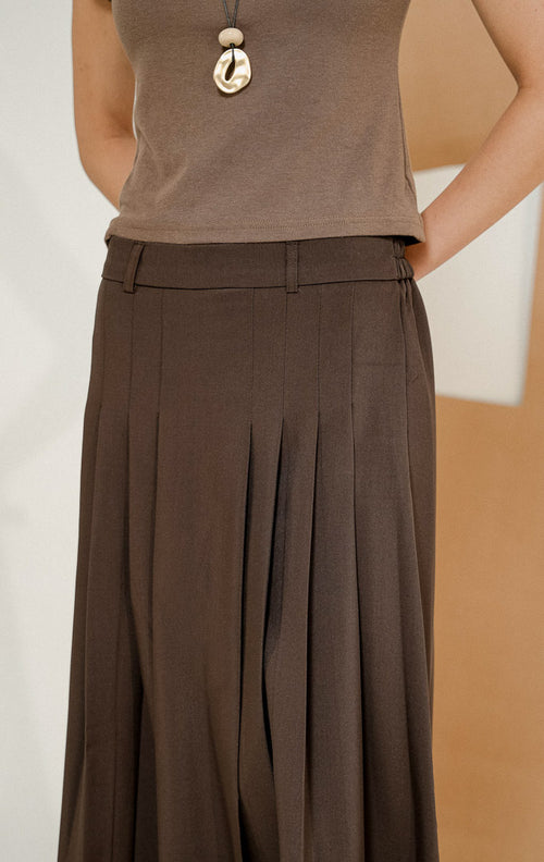 Model berdiri mengenakan Maiden Box Pleats Maxi Skirt Dark Brown, produk rok panjang wanita model maxi warna cokelat tua dengan detail lipit kotak. 