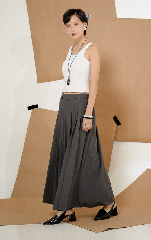 Model berdiri mengenakan Maiden Box Pleats Maxi Skirt Dark Grey, produk rok panjang wanita model maxi warna abu-abu tua dengan detail lipit kotak dipadukan tank top putih.