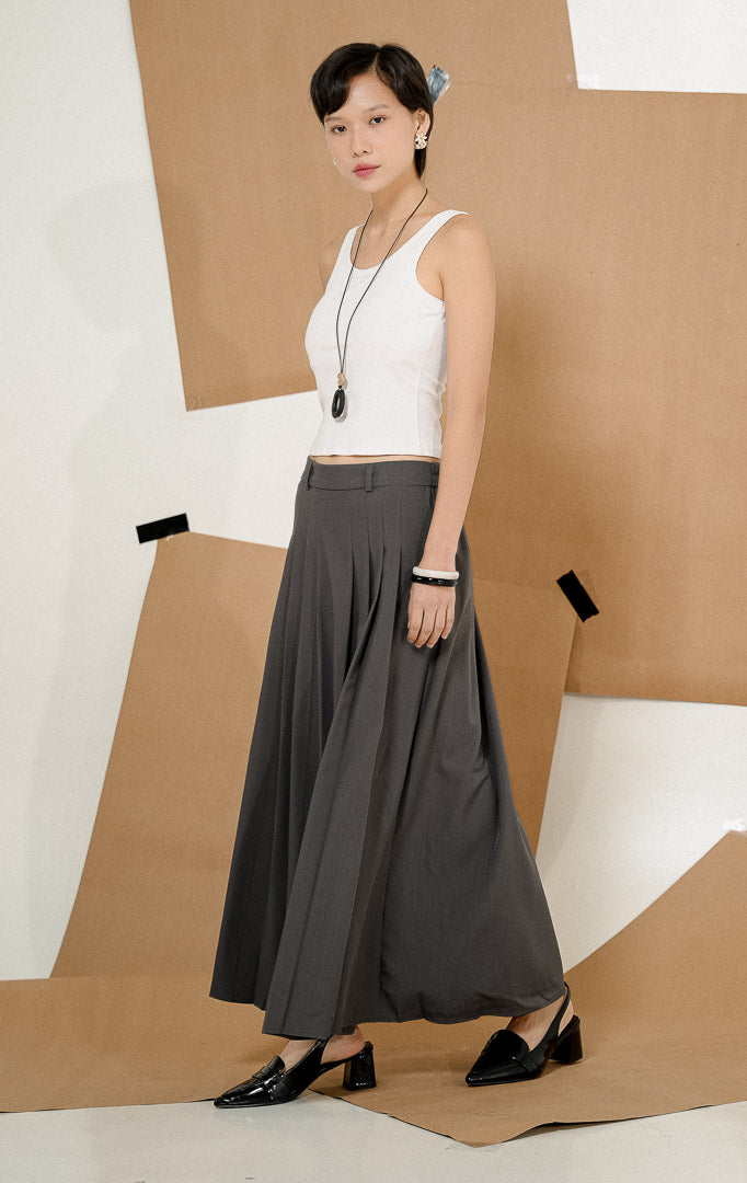 Model berdiri mengenakan Maiden Box Pleats Maxi Skirt Dark Grey, produk rok panjang wanita model maxi warna abu-abu tua dengan detail lipit kotak dipadukan tank top putih.