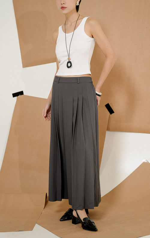 Model berdiri mengenakan Maiden Box Pleats Maxi Skirt Dark Grey, produk rok panjang wanita model maxi warna abu-abu tua dengan detail lipit kotak dipadukan tank top putih.