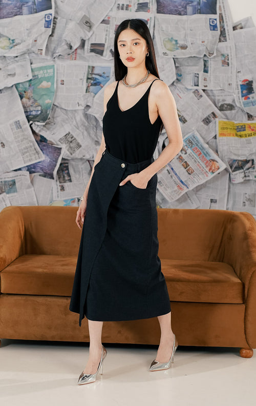 Wanita mengenakan Marjorie Skirt warna navy blue, rok midi panjang dengan model wrap asimetris, detail kancing depan, dan saku samping.