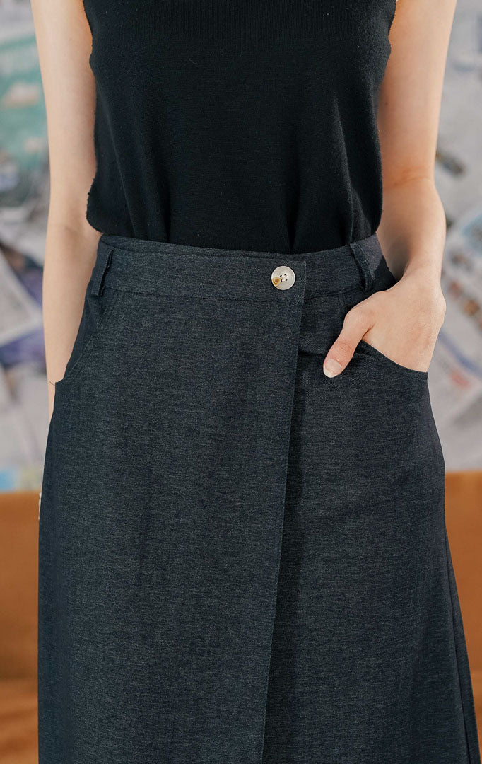Wanita mengenakan Marjorie Skirt warna navy blue, rok midi panjang dengan model wrap asimetris, detail kancing depan, dan saku samping.1