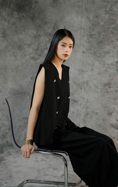 Atasan rompi wanita Marzia warna hitam tanpa lengan dengan potongan leher v-neck, dilengkapi detail aksen payet mewah dan bros bentuk bunga yang manis. 