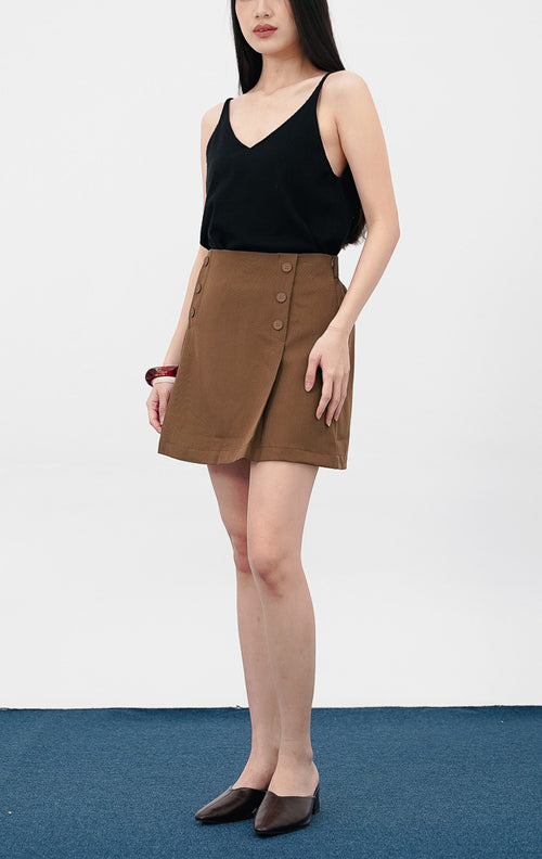 Model mengenakan Matteo Skort warna dark brown, rok celana pendek dengan detail deretan kancing dekoratif di sisi depan dan potongan pinggang tinggi (high-waist).