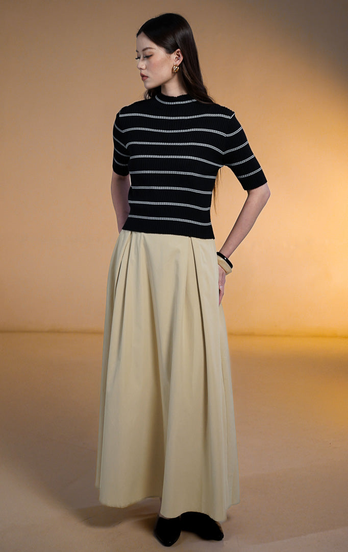 Model wanita mengenakan Mervin High Neck Stripe Knit Top warna hitam, atasan rajut lengan pendek motif garis putih horizontal dengan kerah tinggi (mock neck) bertepi gelombang, dipadukan dengan rok panjang krem