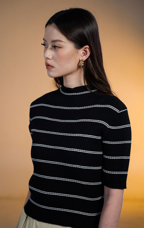 Model wanita mengenakan Mervin High Neck Stripe Knit Top warna hitam, atasan rajut lengan pendek motif garis putih horizontal dengan kerah tinggi (mock neck) bertepi gelombang, dipadukan dengan rok panjang krem