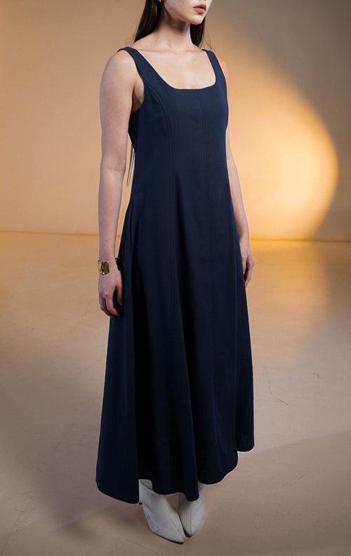 Model wanita mengenakan Miren Midi Dress warna biru dongker (navy), gaun terusan potongan A-line selutut dengan kerah kotak (square neck), bahan katun tegas, dan saku di sisi samping