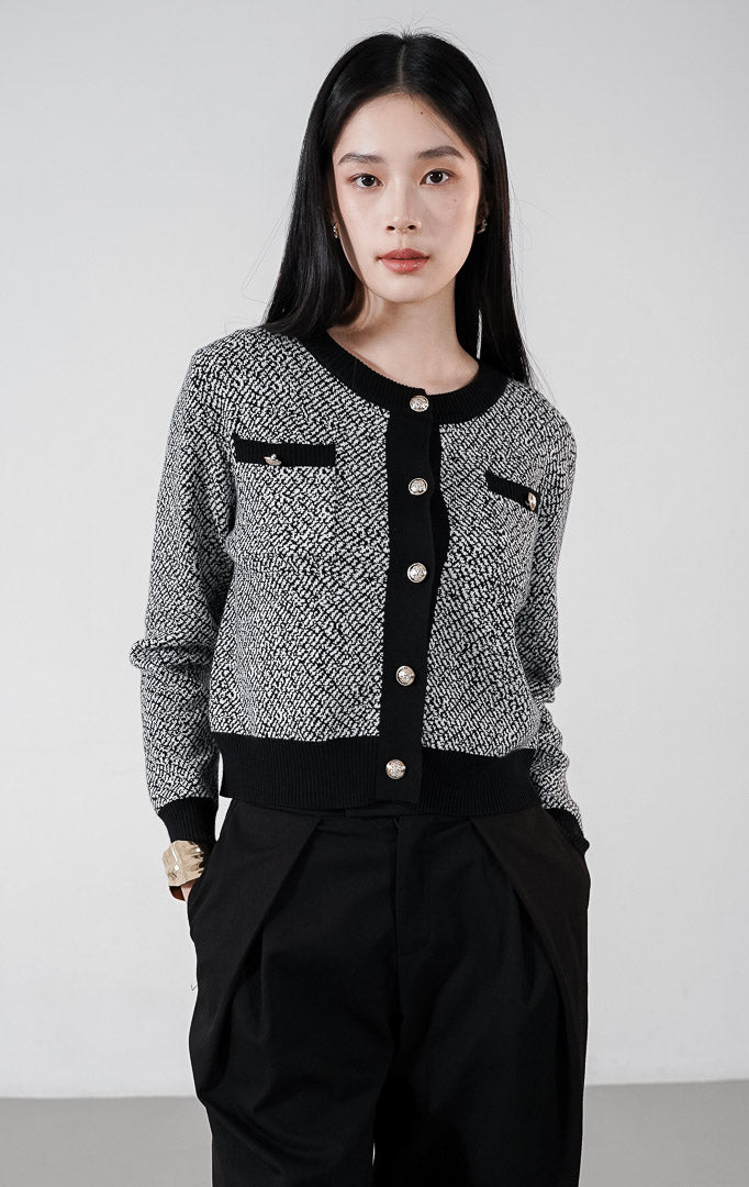 Model mengenakan Monique Cardigan warna black, kardigan rajut pola dua warna dengan detail garis hitam kontras, dua saku dada, dan kancing emas bulat di bagian depan.