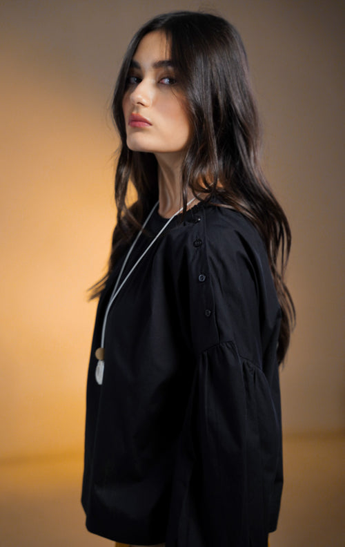 Model wanita mengenakan Morgana Puffy Sleeve Cropped Top warna hitam, atasan lengan panjang balon (puffy) dengan detail deretan kancing di sepanjang garis bahu hingga lengan, dipadukan dengan kalung panjang