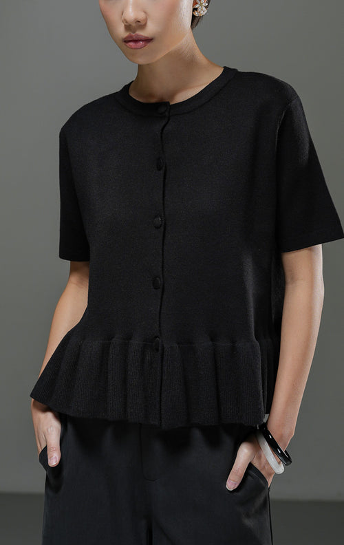 Atasan rajut wanita Naari Top warna hitam lengan pendek dengan kerah bulat, kancing depan bungkus, dan detail rumbai (ruffle hem) rajut di bagian bawah. 