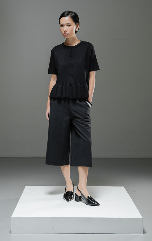 Atasan rajut wanita Naari Top warna hitam lengan pendek dengan kerah bulat, kancing depan bungkus, dan detail rumbai (ruffle hem) rajut di bagian bawah. 