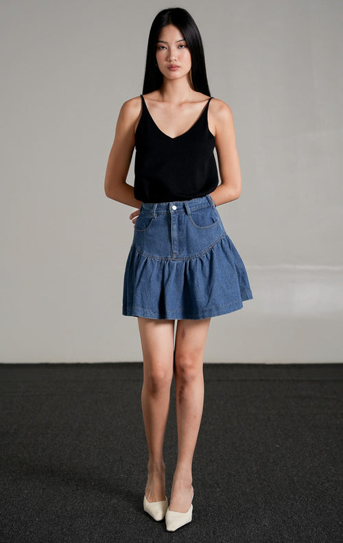 Nathalia Skirt Indigo, rok mini jeans warna biru tua dengan detail lipatan ruffle di bagian bawah, dipadukan dengan tank top hitam.