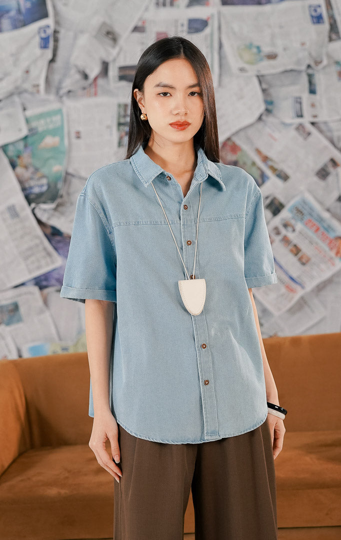 Model wanita memakai Nathaniel Top Denim, kemeja atasan bahan denim ringan warna biru muda lengan pendek dengan kancing depan coklat dan potongan loose fit.1