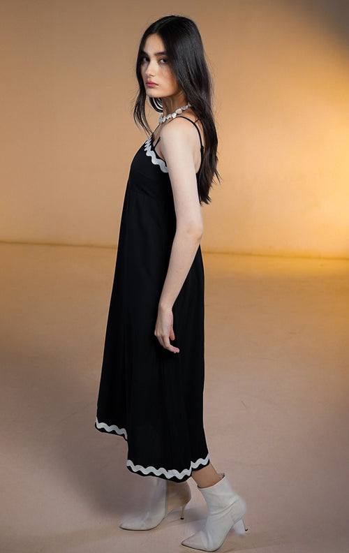 Model wanita mengenakan Nolene Dress Black, gaun midi hitam model tali spageti (cami) dengan pinggiran bergelombang putih (scalloped trim) di leher dan bawah, dipadukan dengan sepatu boots putih