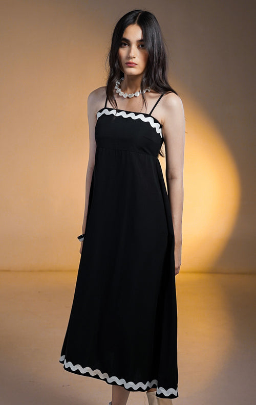 Model wanita mengenakan Nolene Dress Black, gaun midi hitam model tali spageti (cami) dengan pinggiran bergelombang putih (scalloped trim) di leher dan bawah, dipadukan dengan sepatu boots putih