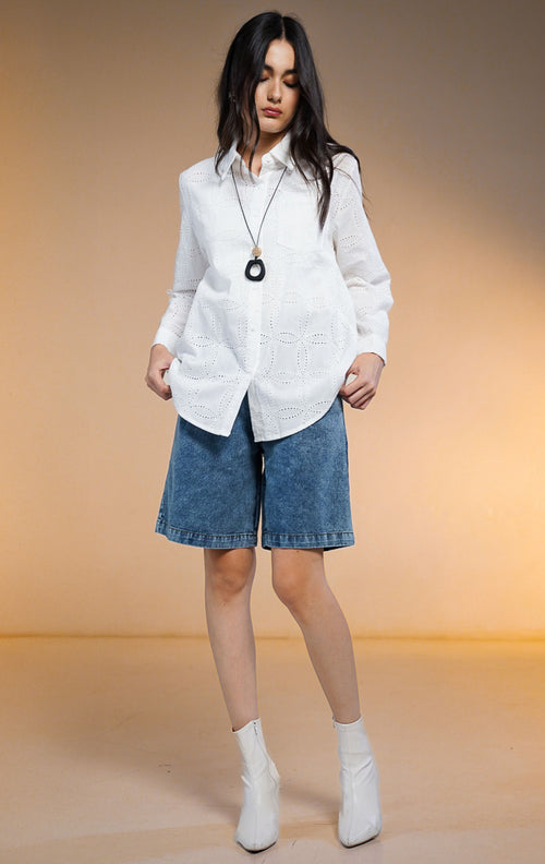 Model wanita mengenakan Nome Floral Embroidered Eyelet Shirt warna putih, kemeja lengan panjang kancing depan dengan motif bordir lubang (eyelet) dan saku dada, dipadukan dengan celana pendek denim dan sepatu boots putih