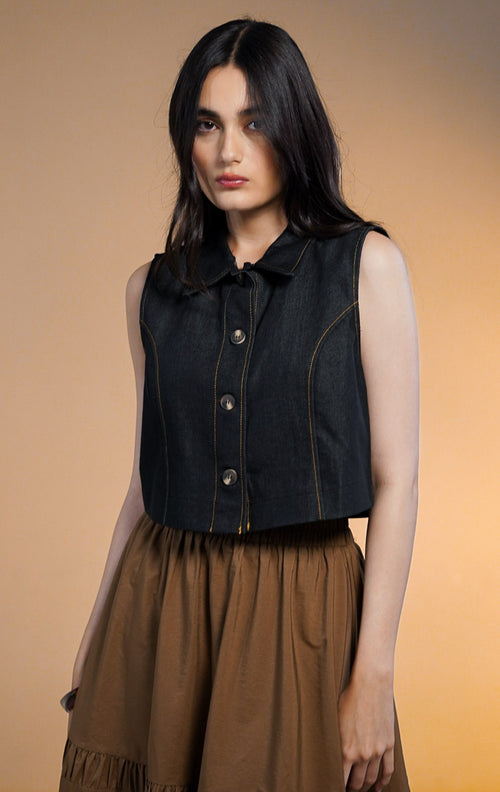 Model wanita mengenakan Norani Sleeveless Crop Denim Top, atasan rompi denim biru tua berkerah dengan kancing depan dan jahitan benang kuning kontras, dipadukan dengan rok rimpel panjang warna cokela