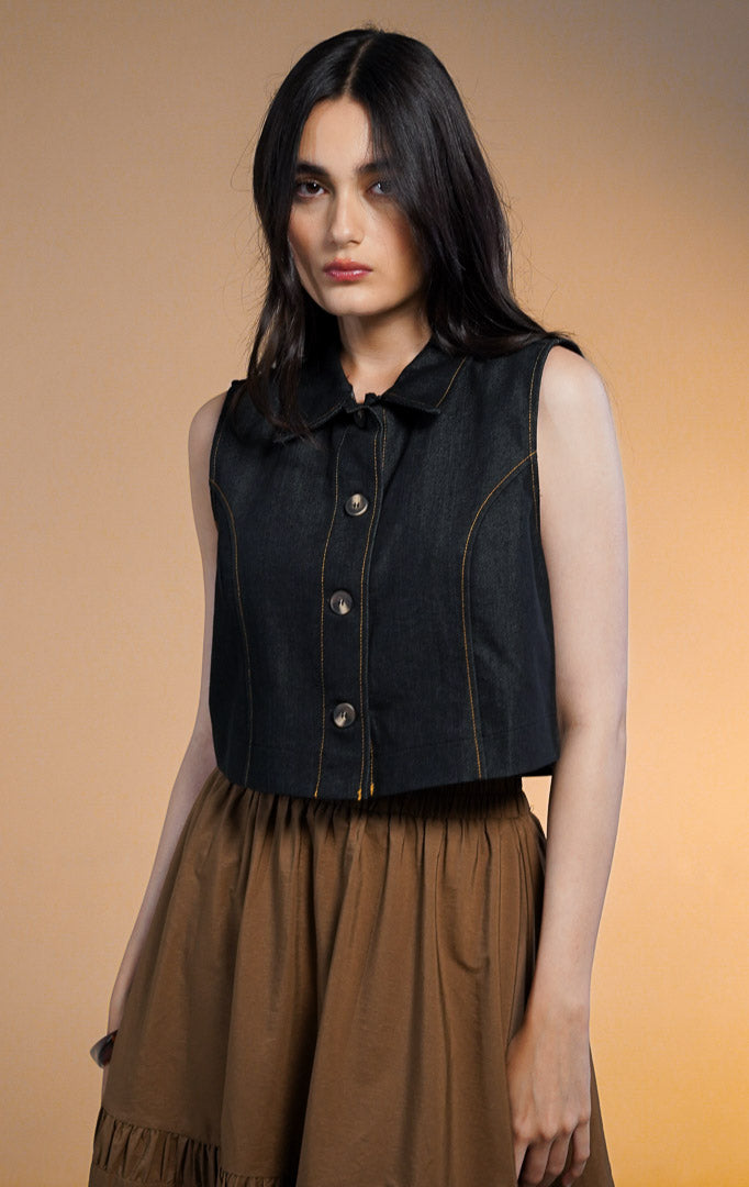 Model wanita mengenakan Norani Sleeveless Crop Denim Top, atasan rompi denim biru tua berkerah dengan kancing depan dan jahitan benang kuning kontras, dipadukan dengan rok rimpel panjang warna cokela