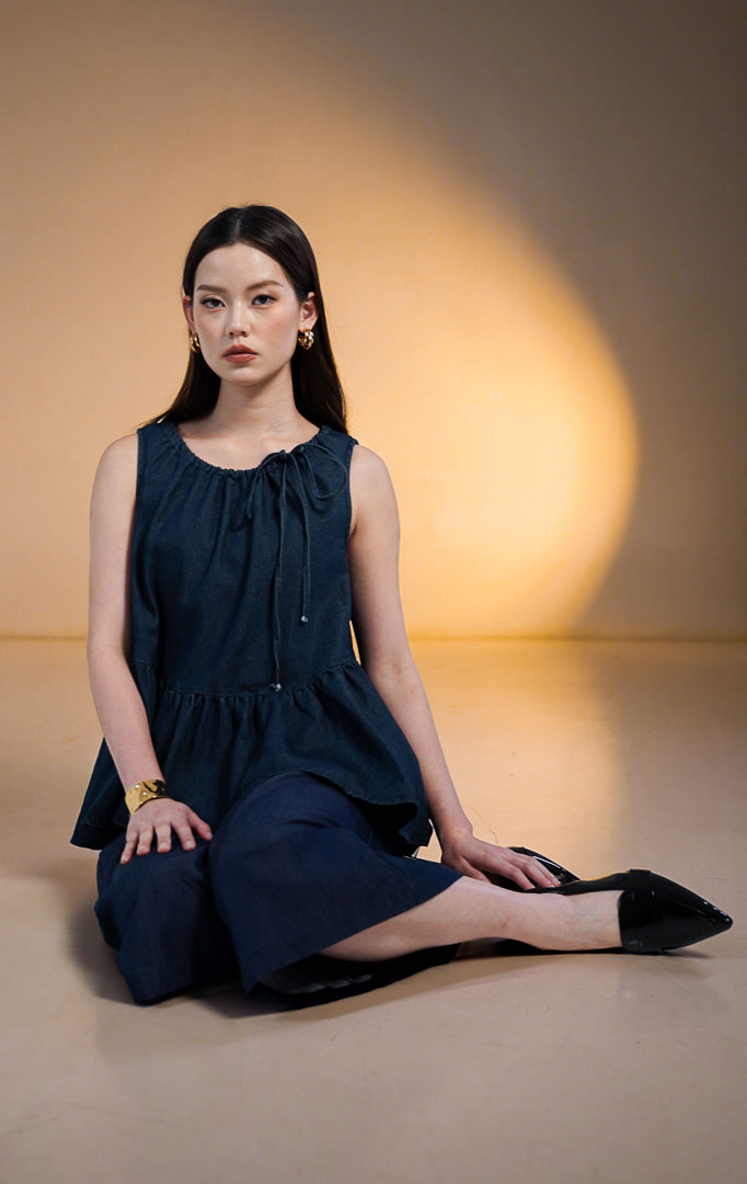 Model wanita mengenakan Norani Sleeveless Crop Denim Top, atasan denim biru tua tanpa lengan dengan kerah kerut tali serut (drawstring tie) dan ujung bawah model ruffle mekar (peplum), dipadukan dengan celana kulot denim senada