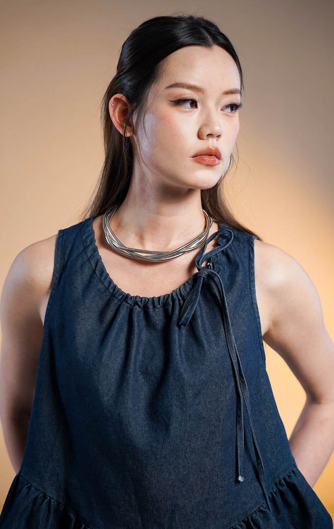 Model wanita mengenakan Norani Sleeveless Crop Denim Top, atasan denim biru tua tanpa lengan dengan kerah kerut tali serut (drawstring tie) dan ujung bawah model ruffle mekar (peplum), dipadukan dengan celana kulot denim senada