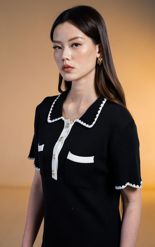 Model wanita mengenakan Perine Crochet Trim Mini Knit Dress warna hitam, gaun rajut lengan pendek dengan kerah dan saku dada bertepi renda putih (crochet trim) serta kancing depan emas.