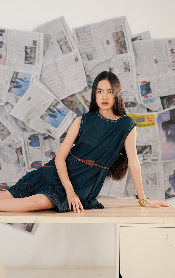 Model wanita memakai Piper Dress Navy, dress denim tanpa lengan warna biru tua dengan detail jahitan kontras (contrast stitching) di leher dan aksen ruffle di bagian bawah rok.1