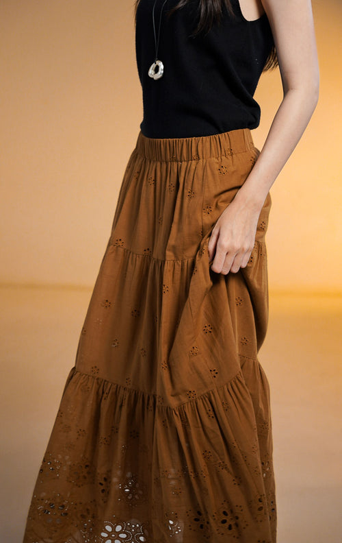 Model wanita mengenakan Primrose Tiered Midi Skirt warna cokelat, rok midi model susun (tiered) dengan bordir lubang bunga kecil (eyelet) dan pinggang karet, dipadukan dengan atasan tanpa lengan hitam
