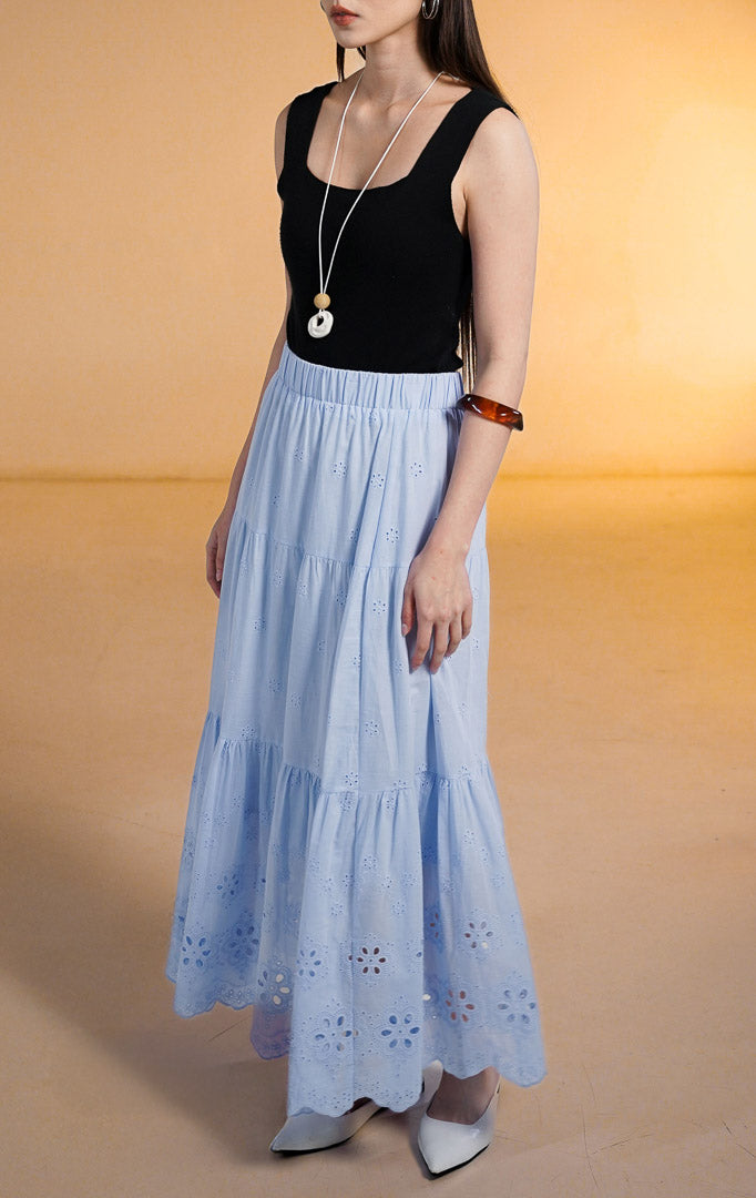 Model wanita mengenakan Primrose Tiered Skirt warna biru muda (baby blue), rok susun panjang dengan motif bordir lubang (eyelet) dan pinggang karet, dipadukan dengan tank top hitam dan sepatu putih.