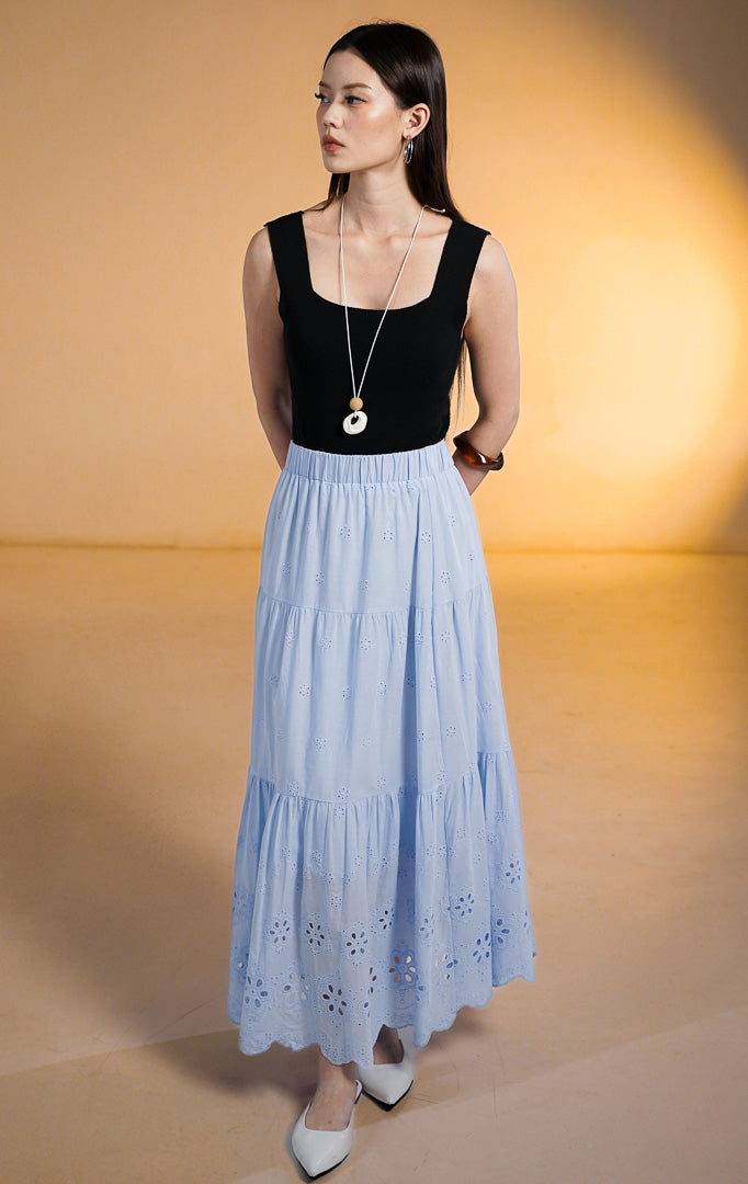 Model wanita mengenakan Primrose Tiered Skirt warna biru muda (baby blue), rok susun panjang dengan motif bordir lubang (eyelet) dan pinggang karet, dipadukan dengan tank top hitam dan sepatu putih.