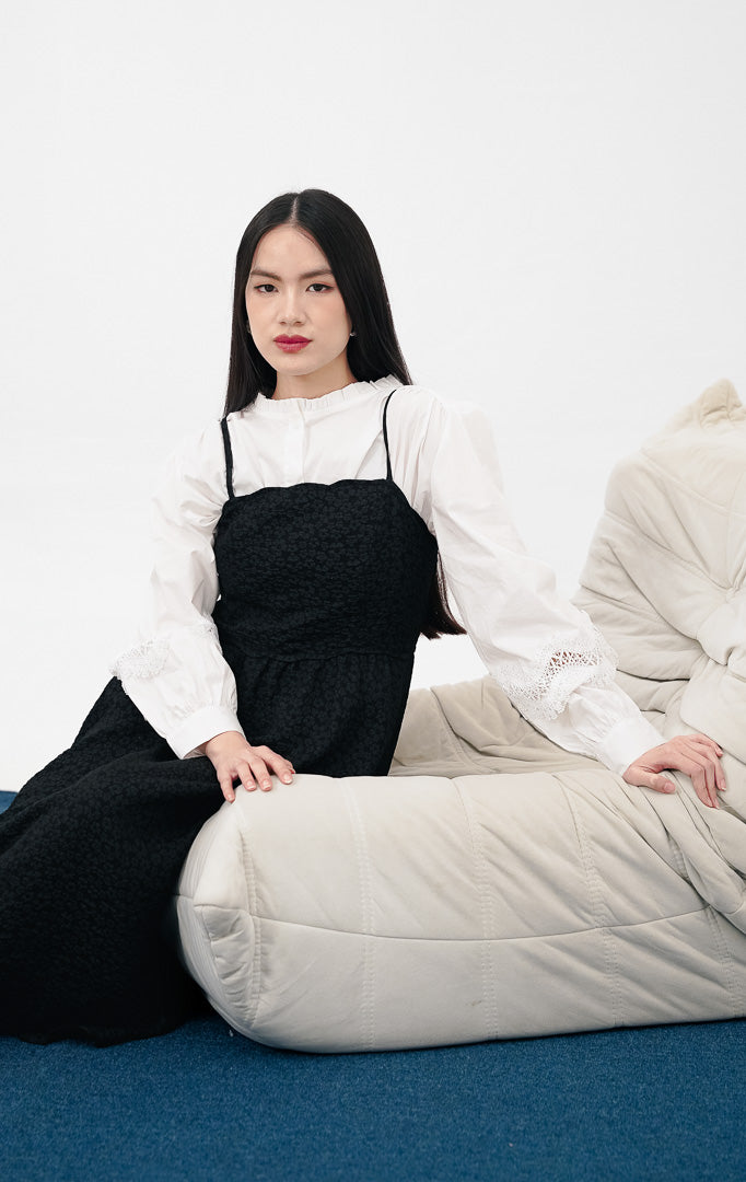  Model mengenakan Raelyn Dress warna black sebagai luaran, gaun maxi tali bahan jacquard tekstur bunga kecil dipadukan dengan kemeja Princeton putih di bagian dalam.