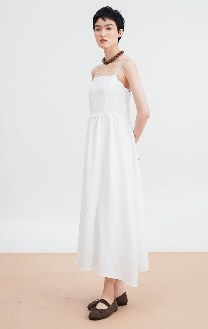 Model mengenakan Raelyn Dress warna white, gaun maxi tali bahan jacquard dengan tekstur pola halus, siluet A-line, dipadukan dengan kalung cokelat besar.