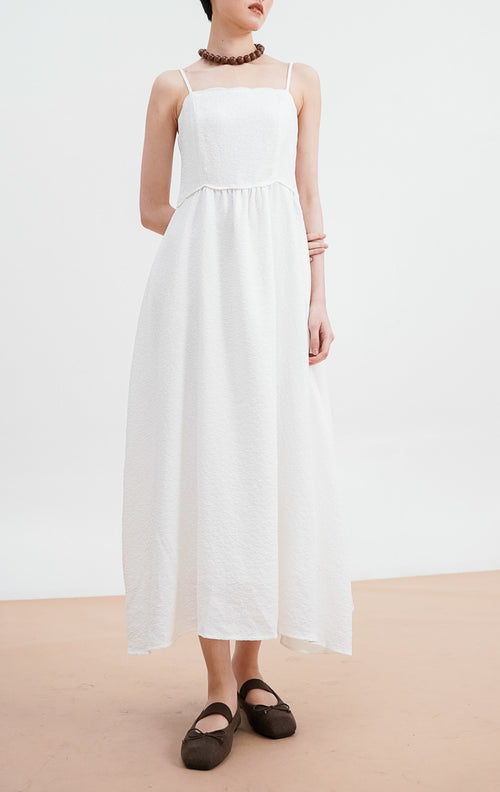 Model mengenakan Raelyn Dress warna white, gaun maxi tali bahan jacquard dengan tekstur pola halus, siluet A-line, dipadukan dengan kalung cokelat besar.