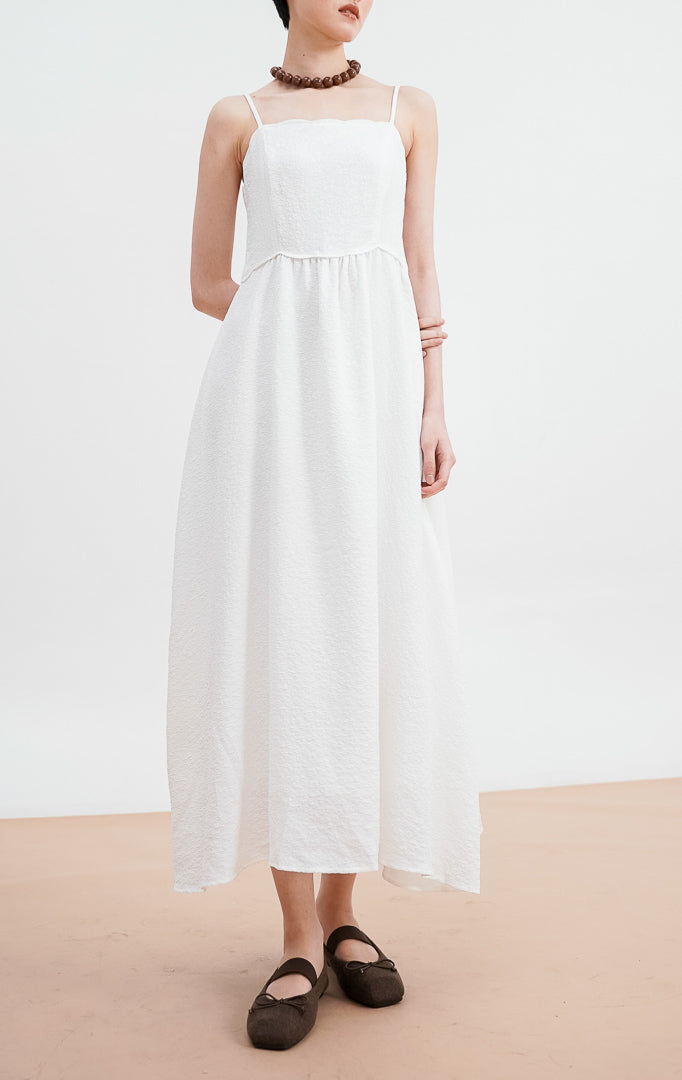 Model mengenakan Raelyn Dress warna white, gaun maxi tali bahan jacquard dengan tekstur pola halus, siluet A-line, dipadukan dengan kalung cokelat besar.