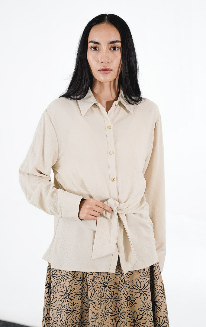 Raphina Top warna ivory, kemeja lengan panjang bertekstur crinkle dengan detail tali pita yang diikat di bagian pinggang dan kancing emas.