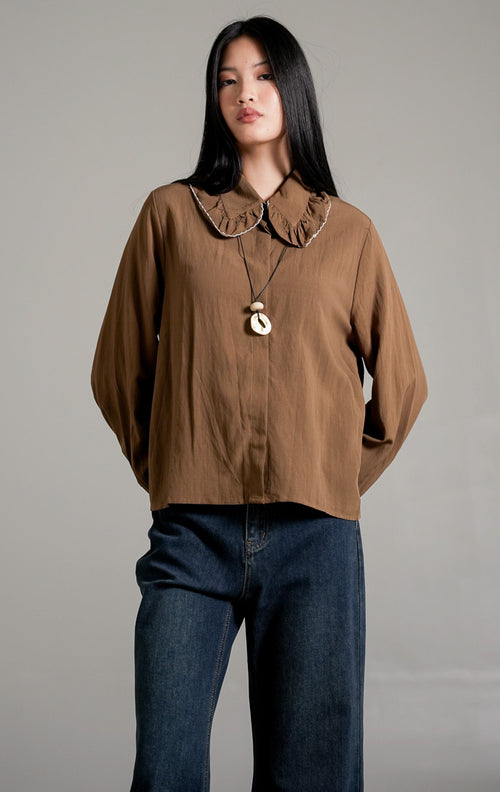 Rebecca Top Brown, kemeja katun lengan panjang warna cokelat dengan detail kerah scallop lebar, dipadukan dengan celana jeans biru tua.