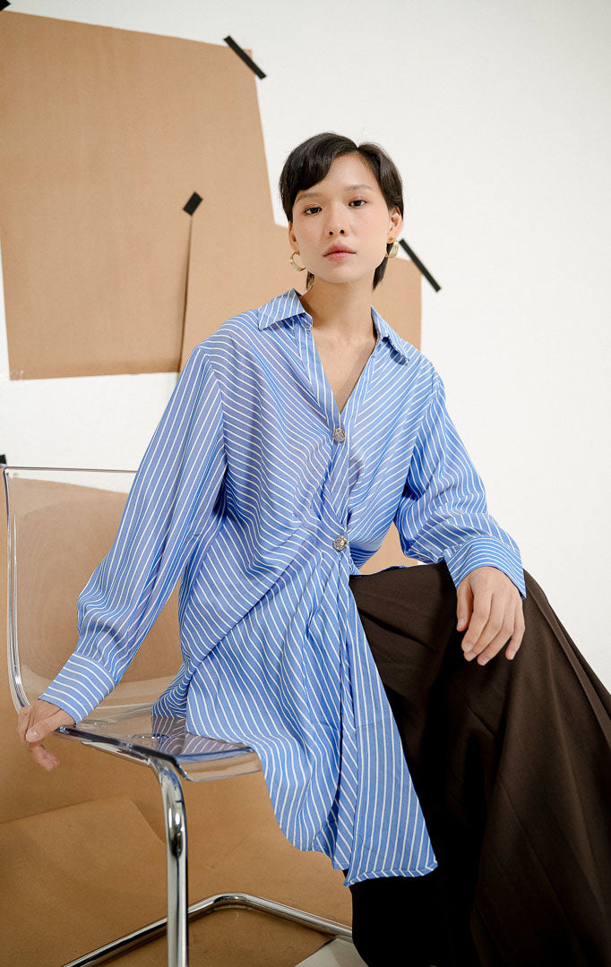 Model duduk di kursi mengenakan Rhiannon Drape Striped Blouse Blue, produk kemeja salur wanita lengan panjang warna biru-putih dengan detail drape kerutan di depan dan kancing emas. 