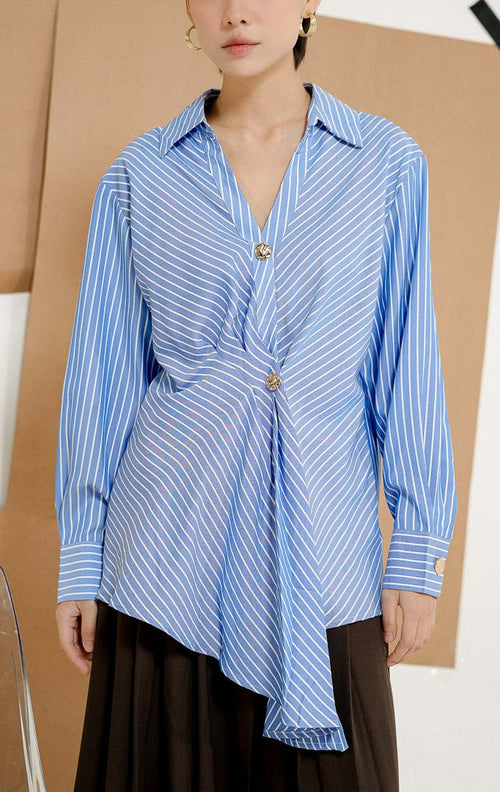 Model duduk di kursi mengenakan Rhiannon Drape Striped Blouse Blue, produk kemeja salur wanita lengan panjang warna biru-putih dengan detail drape kerutan di depan dan kancing emas. 
