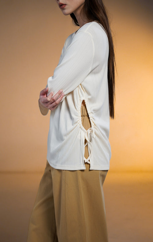 Model wanita mengenakan Rinata Long Sleeve Top warna putih gading (ivory), atasan lengan panjang bertekstur rib halus dengan detail tali serut (drawstring) dan lubang di sisi pinggang kiri, dipadukan dengan celana panjang cokelat mustard