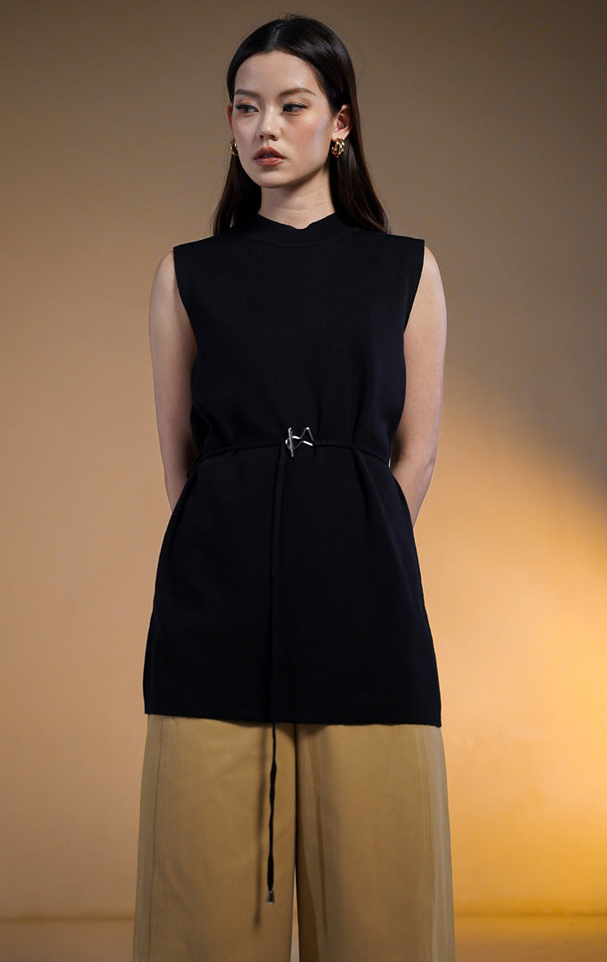Model wanita mengenakan Rivane Sleeveless Knit Long Top warna hitam, atasan tunik rajut panjang tanpa lengan dengan kerah tinggi dan ikat pinggang kecil bergesper perak unik, dipadukan dengan celana kulot krem