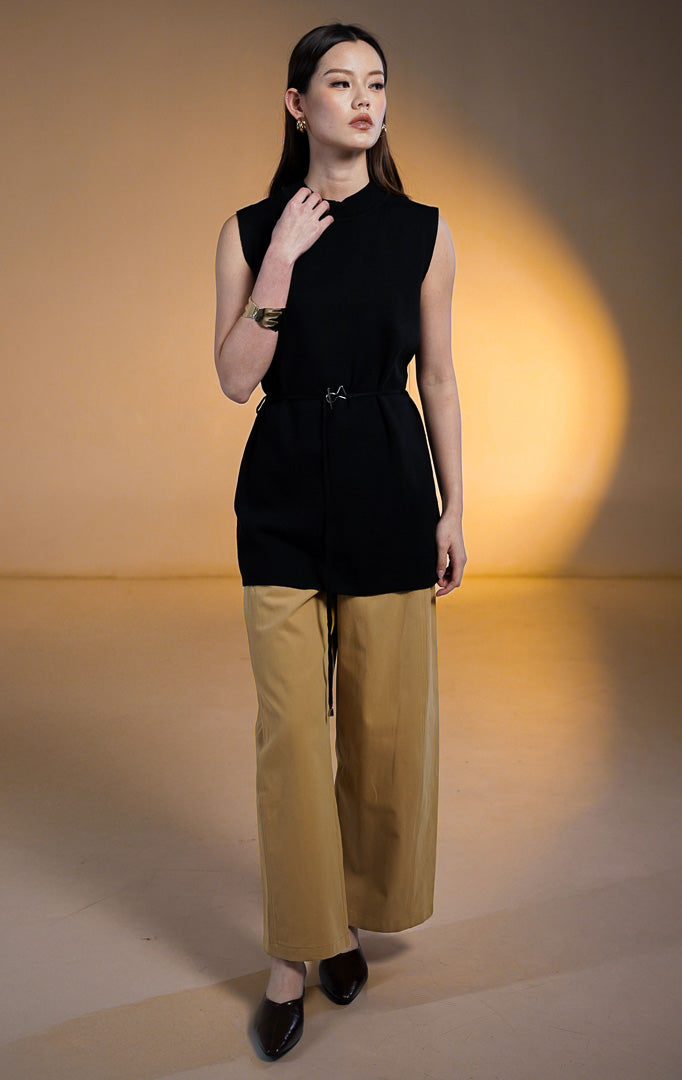 Model wanita mengenakan Rivane Sleeveless Knit Long Top warna hitam, atasan tunik rajut panjang tanpa lengan dengan kerah tinggi dan ikat pinggang kecil bergesper perak unik, dipadukan dengan celana kulot krem