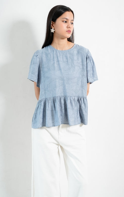 Atasan wanita lengan pendek warna biru pastel (dusty blue) Rivena Top berbahan tekstur crinkle ringan (lightweight crinkle texture) dengan kerah bulat (round neckline) dan detail bagian bawah bertingkat kerut (softly gathered tiered hem). 

