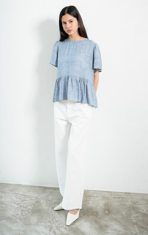Atasan wanita lengan pendek warna biru pastel (dusty blue) Rivena Top berbahan tekstur crinkle ringan (lightweight crinkle texture) dengan kerah bulat (round neckline) dan detail bagian bawah bertingkat kerut (softly gathered tiered hem). 

