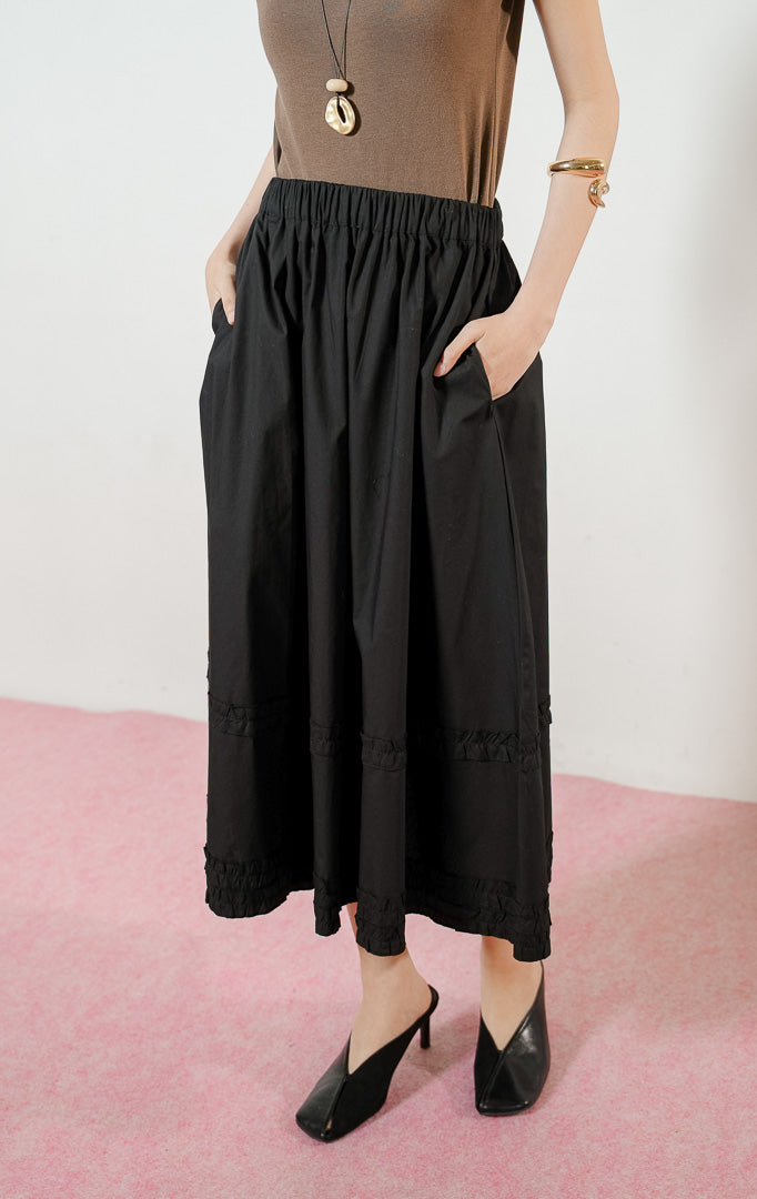 Model wanita mengenakan Arabella Skirt warna black, rok midi panjang wanita berwarna hitam dengan pinggang karet dan detail aksen kerut ruffle horizontal.