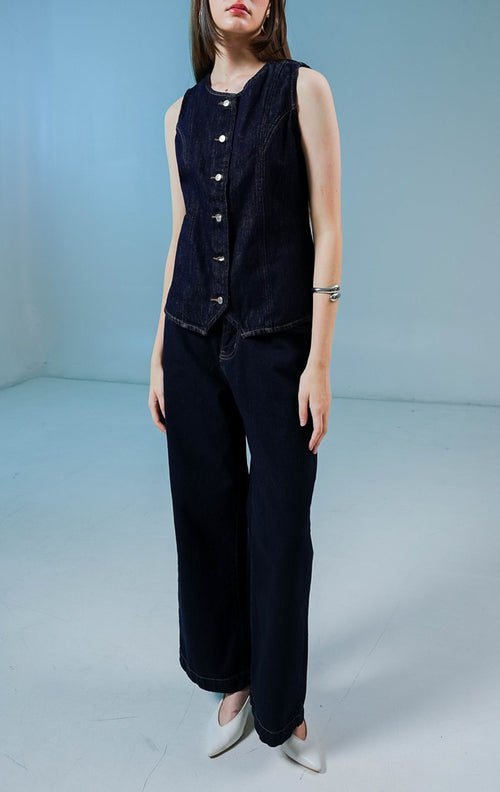 Model wanita mengenakan Vior Button-Up Vest warna dark denim, atasan rompi vest denim wanita berwarna biru dongker gelap dengan detail kancing depan logam. 