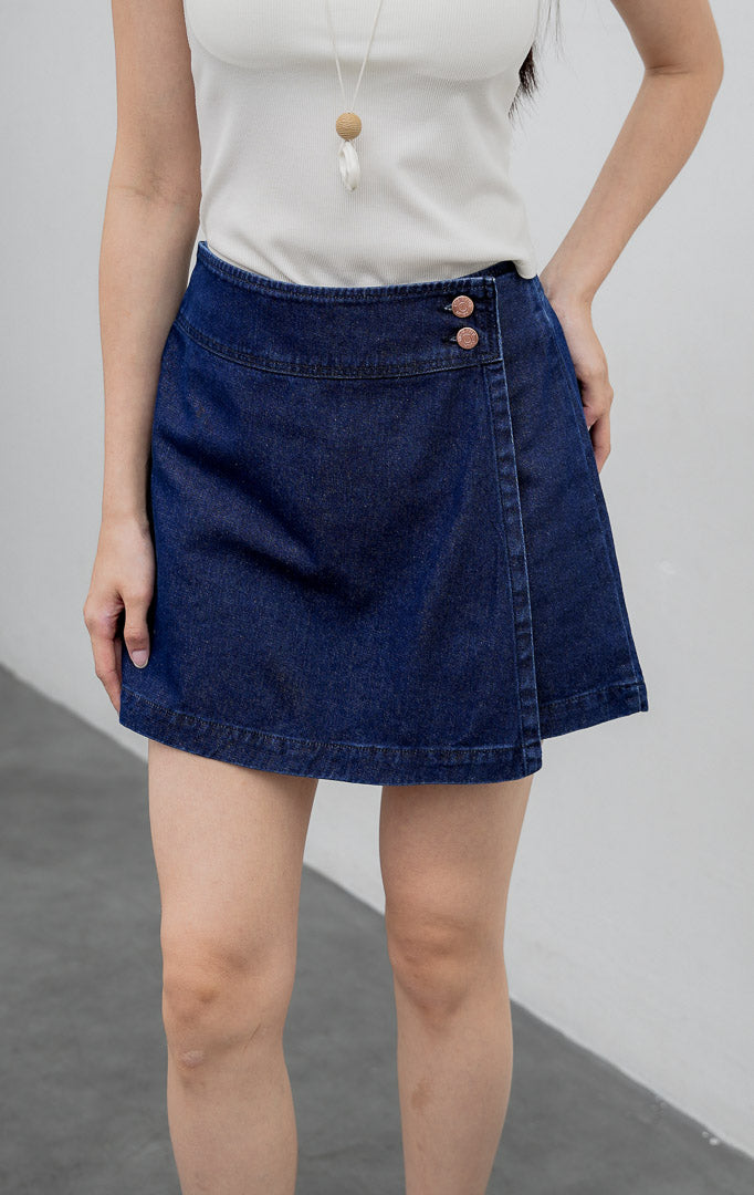 Rok celana (skort) wanita berbahan denim warna biru tua (dark denim) Ronan Skort dengan potongan asimetris di bagian depan, kancing samping tembaga, dan saku belakang.