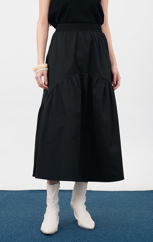Model mengenakan Rosalia Skirt warna black, rok panjang dengan detail pinggang elastis dan potongan bertingkat (tiered) yang melebar di bagian bawah.