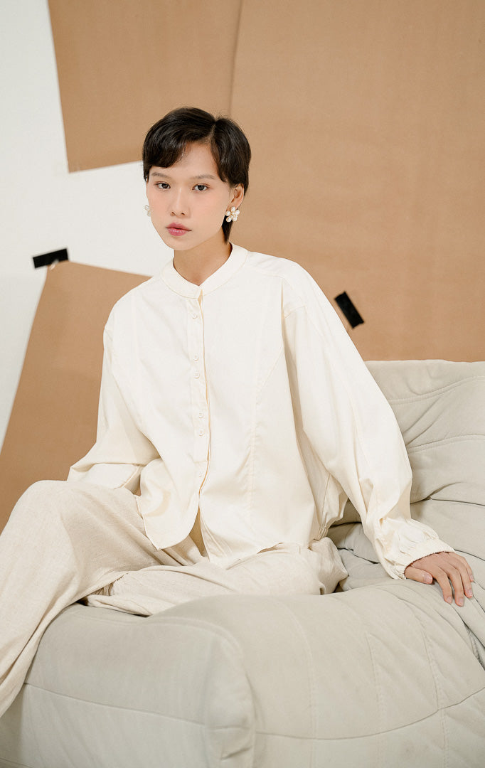 Model duduk di sofa beanbag mengenakan Rosemarie Top Ivory, produk atasan blouse kemeja wanita lengan panjang warna putih gading dengan detail kancing depan. 